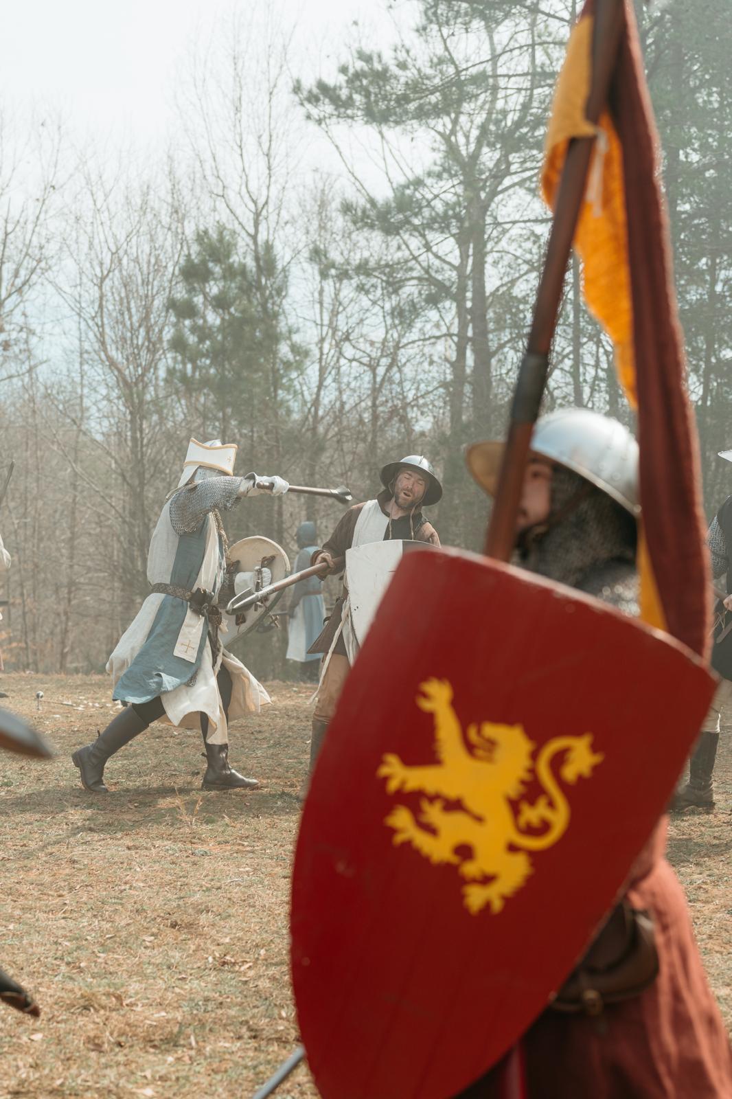 Ludwig, an early 13th-century Flemish mercenary holding a red shield with a lion emblazoned on it and a spear aloft, stands in front of a battle, seemingly oblivious to it